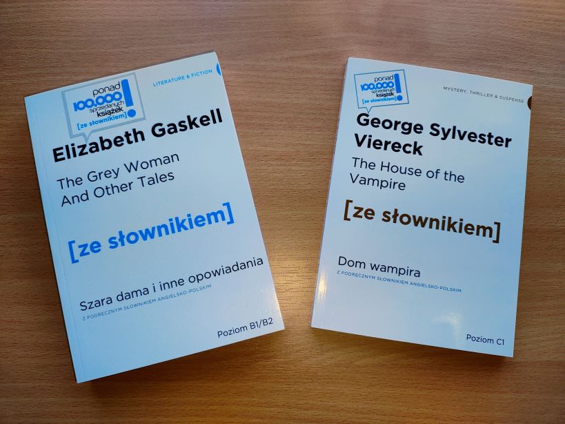 Zdjęcie przedstawia dwie książki, które podarowało wydawnictwo. Mają białe okładki i czarno-niebieskie napisy.  Leżą na drewnianym blacie.